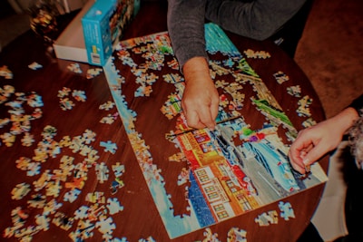 Two people are assembling a colorful jigsaw puzzle on a wooden table. The partially completed puzzle features a scenic image with buildings and a road. Several loose puzzle pieces are scattered around the table, and a puzzle box is placed nearby.