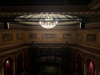 An ornate theater interior featuring a richly decorated ceiling with intricate patterns and a large chandelier as the centerpiece. The walls are adorned with classical paintings and intricate moldings. Dimly lit, the scene conveys a sense of historical elegance and grandeur, highlighting the luxurious design elements typical of classic theater architecture.