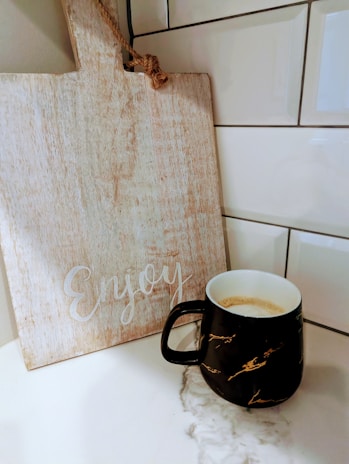 A textured wooden cutting board with the word 'Enjoy' written on it stands upright against a tiled wall. Next to it is a black ceramic mug with a marble-like pattern, filled with coffee, resting on a smooth, light-colored countertop.
