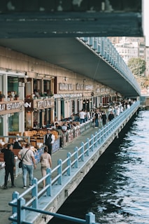 A bustling riverside walkway with numerous people strolling and sitting at outdoor tables of various restaurants. The structure is lined with shops and eateries, their names prominently displayed. The walkway is bordered by a railing with a body of water visible on one side. The atmosphere appears lively and social.