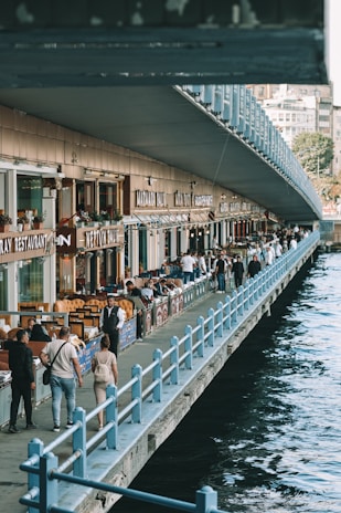 A bustling riverside walkway with numerous people strolling and sitting at outdoor tables of various restaurants. The structure is lined with shops and eateries, their names prominently displayed. The walkway is bordered by a railing with a body of water visible on one side. The atmosphere appears lively and social.
