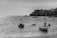 Several small boats are floating on a shimmering body of water, with a rocky coastline visible in the background. Buildings and palm trees line the coastal area, creating a silhouette against the sky.