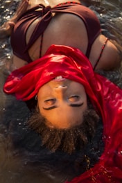A woman is lying in shallow water wearing a dark swimsuit with a vibrant red fabric draped across her. Her eyes are closed, and her hair is wet, blending into the water's reflections.