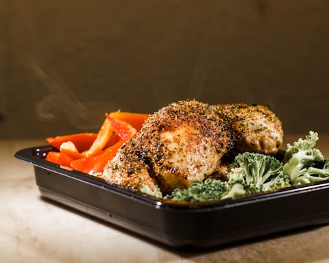 A serving of seasoned grilled chicken is placed in a black container. The chicken is garnished with herbs and spices, accompanied by vibrant red bell pepper slices and fresh broccoli florets. Steam rises from the dish, suggesting it is freshly prepared.