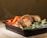 A serving of seasoned grilled chicken is placed in a black container. The chicken is garnished with herbs and spices, accompanied by vibrant red bell pepper slices and fresh broccoli florets. Steam rises from the dish, suggesting it is freshly prepared.