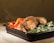 A serving of seasoned grilled chicken is placed in a black container. The chicken is garnished with herbs and spices, accompanied by vibrant red bell pepper slices and fresh broccoli florets. Steam rises from the dish, suggesting it is freshly prepared.