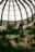 A person stands on a balcony within a large indoor dome structure, characterized by a geometric glass roof. Below, there is an advertisement featuring three smiling women, partially obscured by indoor plants.