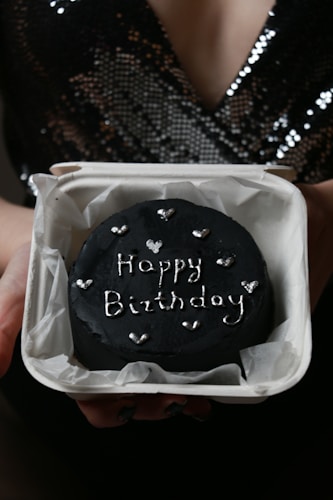 A person is holding a dark chocolate cake inside a white box. The cake has the words 'Happy Birthday' written in silver icing with small hearts decorating it. The background shows the person wearing a black, sequined top.