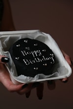 A small, round black cake with 'Happy Birthday' written on it in white icing, adorned with small heart decorations. The cake is resting in a white box lined with parchment paper, held by a hand with black-painted fingernails.