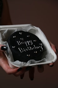 A small, round black cake with 'Happy Birthday' written on it in white icing, adorned with small heart decorations. The cake is resting in a white box lined with parchment paper, held by a hand with black-painted fingernails.