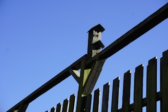 A wooden fence with vertical slats is shown against a clear blue sky. There is a birdhouse mounted on the top of the fence.