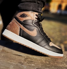 A close-up shot of a stylish sneaker in mid-air, focusing on its detailed stitching and the distinct logo on the side. The sneaker features a combination of black and brown leather with a white rubber sole and visible textural details.