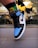 A close-up of a foot wearing a sneaker with black, blue, and white colors, adorned with a prominent Nike swoosh logo. The background features a modern architectural element with reflective surfaces and geometric patterns, creating an urban atmosphere.