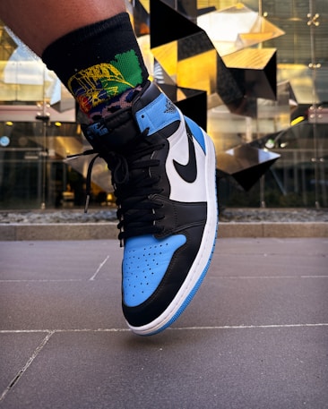 A close-up of a foot wearing a sneaker with black, blue, and white colors, adorned with a prominent Nike swoosh logo. The background features a modern architectural element with reflective surfaces and geometric patterns, creating an urban atmosphere.