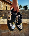 A pair of colorful sneakers with artistic patterns held by a hand, displayed against a stone wall background in an outdoor setting. The shoes have a black base with vibrant multicolored artwork on the fabric and the word 'Supreme' visible on the tongue.