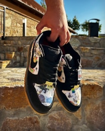 A pair of colorful sneakers with artistic patterns held by a hand, displayed against a stone wall background in an outdoor setting. The shoes have a black base with vibrant multicolored artwork on the fabric and the word 'Supreme' visible on the tongue.
