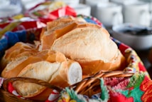 A basket filled with freshly baked bread rolls, with crispy golden crusts and soft white interiors, is placed on a tablecloth with colorful patterns. In the background, there are white cups and bowls, slightly out of focus.