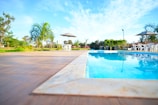 A serene outdoor setting featuring a clear blue swimming pool surrounded by a patio. There are a few white plastic chairs and tables with umbrellas providing shade. Palm trees and a clear blue sky are visible in the background, contributing to a tropical atmosphere.
