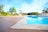A serene outdoor setting featuring a clear blue swimming pool surrounded by a patio. There are a few white plastic chairs and tables with umbrellas providing shade. Palm trees and a clear blue sky are visible in the background, contributing to a tropical atmosphere.