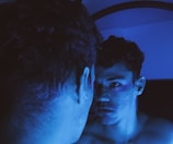 A person looks intensely at their reflection in a mirror, bathed in blue light. There are visible sparkles or glitter on the person's face, adding a mysterious or artistic element.