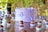 A beautifully decorated purple cake with delicate butterfly decorations and small white sugar pearls on top. The cake is placed on a wooden table surrounded by numerous small chocolate confections, each in a white paper cup. The background features blurred green foliage, creating a natural and serene setting.