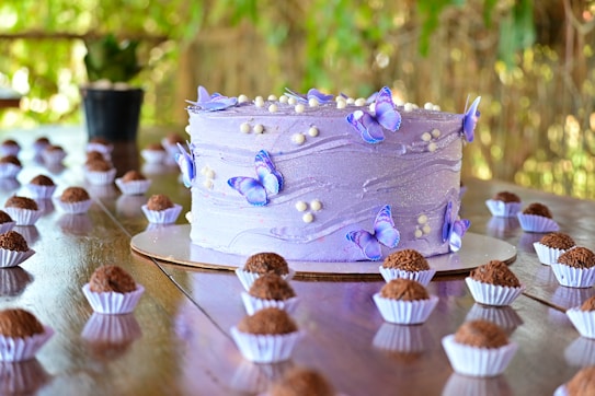 A beautifully decorated purple cake with delicate butterfly decorations and small white sugar pearls on top. The cake is placed on a wooden table surrounded by numerous small chocolate confections, each in a white paper cup. The background features blurred green foliage, creating a natural and serene setting.