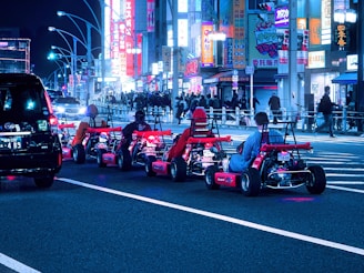 A group of people driving go-karts on a city street at night, surrounded by bright neon lights and bustling activity. The scene is vibrant with various illuminated advertisements and signs in the background. Pedestrians walk along the sidewalk as other vehicles are present on the road.