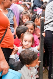 A group of children are gathered closely together, with a young girl at the center looking directly at the camera while holding onto a metal railing. The scene appears crowded, with several children partially visible behind her, and an adult nearby holding a blue bucket.