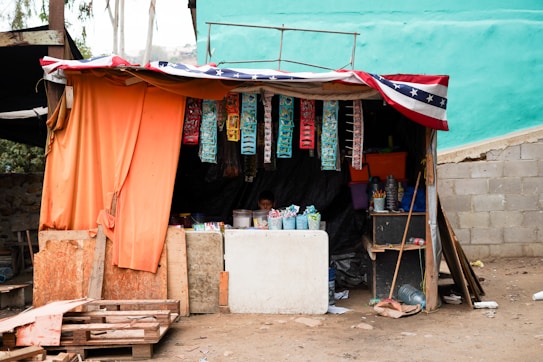 A small makeshift shop with an orange curtain draped over the entrance. Various colorful products, including snacks and household items, hang on display under a canopy that features a pattern resembling the American flag. The shop is situated on a dirt floor with concrete blocks forming part of the exterior walls. Wooden pallets and construction materials are scattered around the area.