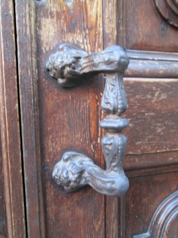 An ornate metallic door handle with intricate designs, mounted on a wooden door. The wood shows signs of age and natural texture, complementing the vintage appearance of the handle.