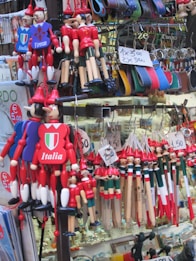 A vibrant market stall displays an assortment of colorful Italian souvenirs, including Pinocchio dolls, keychains, and leather goods. Various small trinkets are neatly arranged with visible price tags.