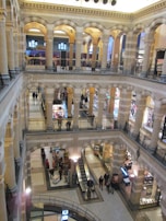 A multi-level indoor space featuring several arched openings and ornate architectural details. The area appears to be a shopping mall with multiple floors accessible via an escalator. People are seen walking or standing on different levels, and various stores are visible, showcasing products and sales.