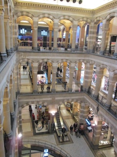A multi-level indoor space featuring several arched openings and ornate architectural details. The area appears to be a shopping mall with multiple floors accessible via an escalator. People are seen walking or standing on different levels, and various stores are visible, showcasing products and sales.