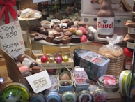 A display of assorted chocolates arranged on a glass stand. The chocolates vary in shape and color, with some wrapped in foil. Beside them are gift tins, a beer bottle, a stack of cookie packages, and a price sign indicating multiple pricing options. The setting suggests a confectionery or chocolate store.