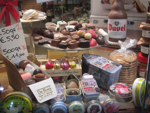 A display of assorted chocolates arranged on a glass stand. The chocolates vary in shape and color, with some wrapped in foil. Beside them are gift tins, a beer bottle, a stack of cookie packages, and a price sign indicating multiple pricing options. The setting suggests a confectionery or chocolate store.