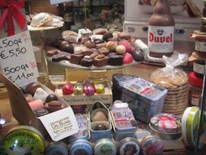 A display of assorted chocolates arranged on a glass stand. The chocolates vary in shape and color, with some wrapped in foil. Beside them are gift tins, a beer bottle, a stack of cookie packages, and a price sign indicating multiple pricing options. The setting suggests a confectionery or chocolate store.