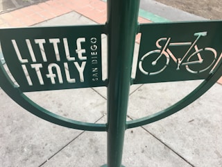 A green metal signpost with cut-out letters and a bicycle symbol. The sign text reads 'Little Italy San Diego'. The pavement beneath the sign is made of concrete tiles with some areas bordered by red bricks.