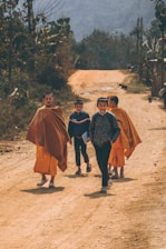 A group of four young boys, two of whom are dressed in traditional orange robes, are walking on a dirt path in a rural setting. The boys appear to be smiling and interacting with each other. The background features a tree-lined path and distant hills, creating a serene and natural atmosphere.