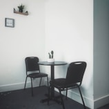 A minimalist corner with a small round table and two black chairs on a gray carpet. A white potted cactus sits in the center of the table. A small shelf with a plant and a framed picture are mounted on the plain white walls.