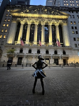 A bronze statue of a confident young girl stands defiantly in front of the grandiose New York Stock Exchange building. The facade is illuminated with bright lights, highlighting its neoclassical columns and American flags. The atmosphere is calm with a few people walking by.