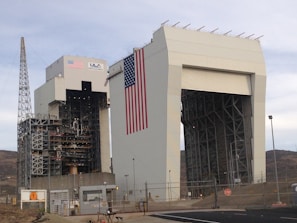 A large industrial structure with intricate metal framework and a massive American flag displayed prominently. The building is labeled with 'ULA' and 'United Launch Alliance' alongside the flag. A tall metal tower stands adjacent, and the area is surrounded by fencing and security measures.