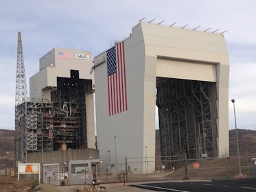 A large industrial structure with intricate metal framework and a massive American flag displayed prominently. The building is labeled with 'ULA' and 'United Launch Alliance' alongside the flag. A tall metal tower stands adjacent, and the area is surrounded by fencing and security measures.