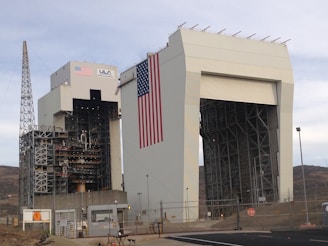 A large industrial structure with intricate metal framework and a massive American flag displayed prominently. The building is labeled with 'ULA' and 'United Launch Alliance' alongside the flag. A tall metal tower stands adjacent, and the area is surrounded by fencing and security measures.