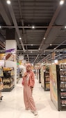 A person stands in the aisle of a grocery store wearing a pink outfit, including a hijab, jacket, and pants, with a matching purse. The shelves are stocked with various products such as bread, chips, and canned goods. The store has a bright, well-lit interior with a modern ceiling and white tiled floor.