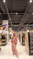 A person stands in the aisle of a grocery store wearing a pink outfit, including a hijab, jacket, and pants, with a matching purse. The shelves are stocked with various products such as bread, chips, and canned goods. The store has a bright, well-lit interior with a modern ceiling and white tiled floor.