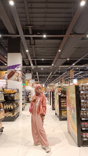 A person stands in the aisle of a grocery store wearing a pink outfit, including a hijab, jacket, and pants, with a matching purse. The shelves are stocked with various products such as bread, chips, and canned goods. The store has a bright, well-lit interior with a modern ceiling and white tiled floor.