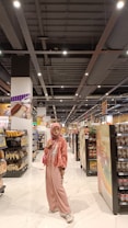 A person stands in the aisle of a grocery store wearing a pink outfit, including a hijab, jacket, and pants, with a matching purse. The shelves are stocked with various products such as bread, chips, and canned goods. The store has a bright, well-lit interior with a modern ceiling and white tiled floor.