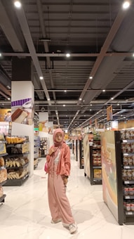 A person stands in the aisle of a grocery store wearing a pink outfit, including a hijab, jacket, and pants, with a matching purse. The shelves are stocked with various products such as bread, chips, and canned goods. The store has a bright, well-lit interior with a modern ceiling and white tiled floor.