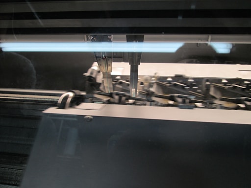 A close-up view of a sewing or knitting machine with metal needles and mechanical components visible under a protective glass cover. The machinery appears to be in use, indicated by the movement of parts and threads.