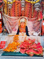 A vibrant and ornate statue of a deity adorned with a crown, surrounded by colorful fabric and garlands of marigold flowers. The background features an intricate, patterned orange and gold cloth. Offerings and religious items are placed in front, including flowers and a red booklet.